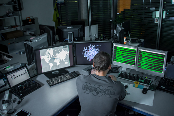 A person sits at a desk facing five computer monitors displaying maps, data, and code.