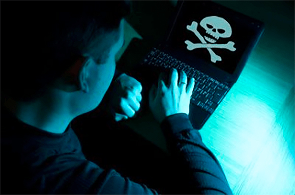 A person in a dark room typing on a laptop that shows a white skull and crossbones symbol on the screen.