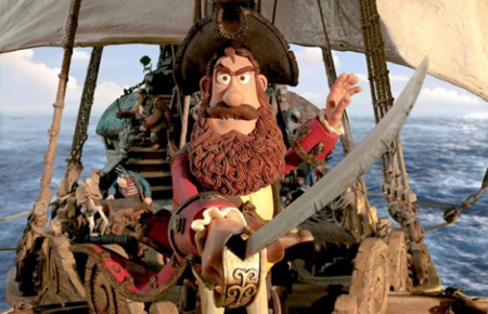 A claymation pirate with a brown beard and large hat holds a curved sword on a sailing ship at sea.