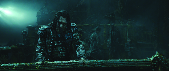 A man with long dark hair and a weathered coat leans over a wooden railing in a dark, misty environment.