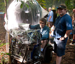 A man sits behind electronic equipment covered in plastic while another man stands nearby in a forest.