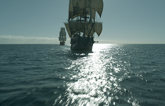 Two large sailing ships with full sails traveling across a shimmering, sunlit ocean under a clear sky.