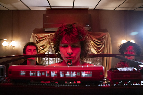 Three men look forward from behind a red model train set in a room with warm lighting.