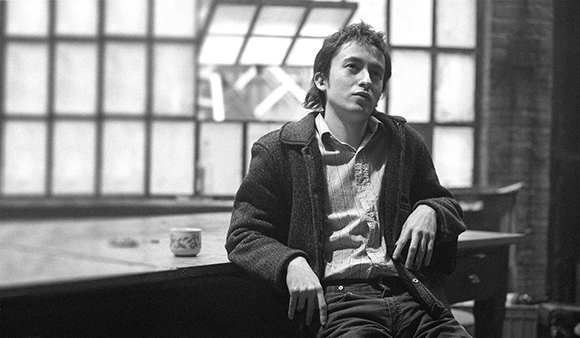Black and white portrait of a man sitting on a wooden table in front of large windows.