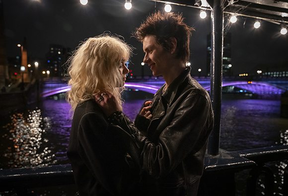A blonde woman and a man with dark spiky hair embrace on a bridge overlooking a purple-lit river at night.