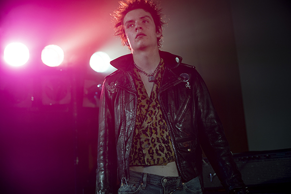 A young man with spiky hair wears a black leather jacket and leopard print shirt under pink stage lights.