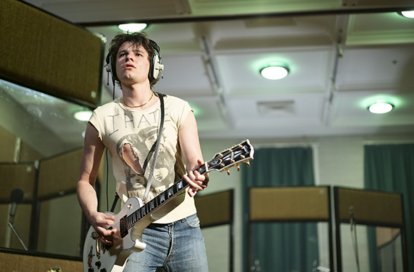 A young man wearing headphones plays a white electric guitar in a recording studio.