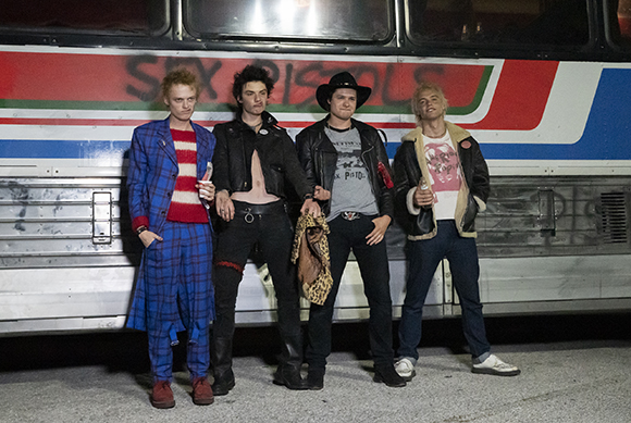 Four men in punk and western-inspired clothing stand in front of a bus with red and blue stripes.