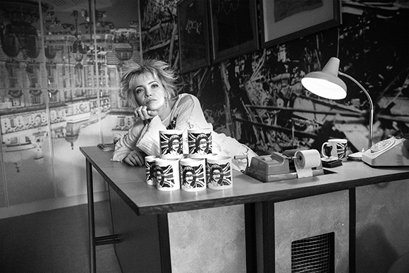 A woman with messy hair leans on a desk lined with several mugs featuring a portrait.