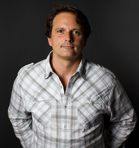 A man with dark hair wearing a white and grey plaid button-down shirt against a dark background.