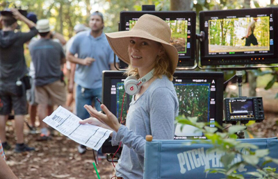 A woman in a wide-brimmed straw hat holds a script and gestures near film monitors in a wooded area.