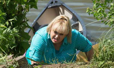 A blonde woman in a turquoise shirt crawls through green foliage near a canoe on the water.