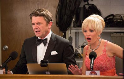A man in a black tuxedo and a woman in a pink sequined dress look shocked behind a desk with microphones.