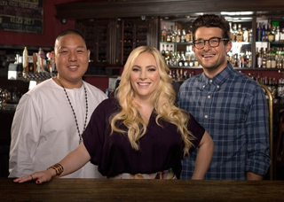 A man in white, a blonde woman in purple, and a man in a blue plaid shirt smiling behind a bar.