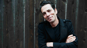 A man with dark hair wearing a black jacket leans against a dark wooden fence.