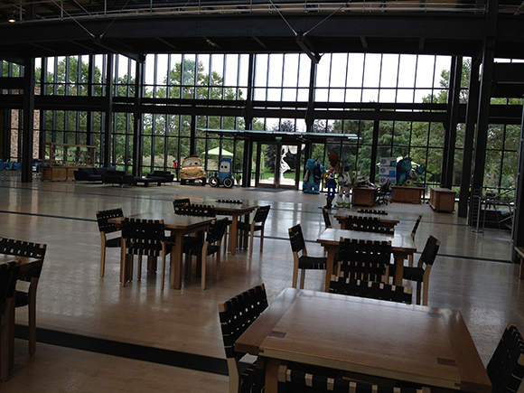 Empty dining hall with wooden tables, chairs, and large windows overlooking trees and character statues.