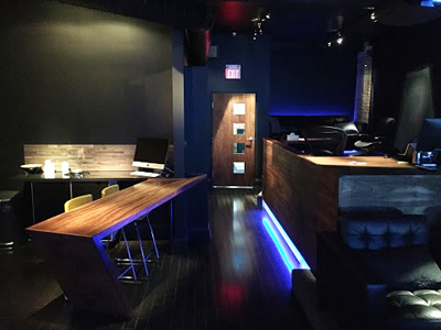 A dark lounge featuring a long wooden bar, high stools, and blue accent lighting along the floor.