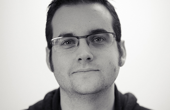 Black and white close-up portrait of a man with short dark hair and rectangular glasses looking forward.