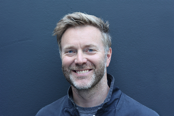 A smiling man with short blond hair and a salt-and-pepper beard against a dark blue background.