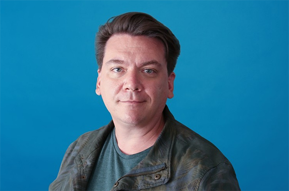 A man with brown hair wearing a camouflage jacket against a solid blue background.