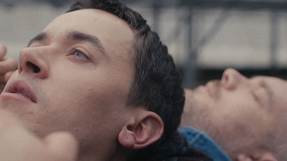 Close-up of two men lying down with their eyes closed or looking upward against a blurred background.