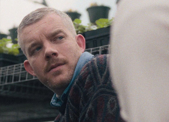 A man with short light hair and light stubble looks toward the side in a soft-focus outdoor setting.
