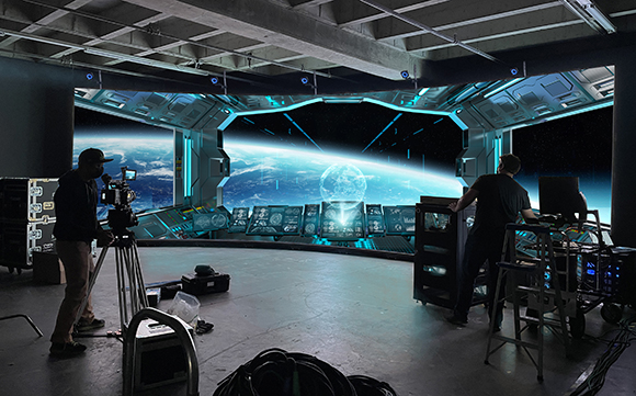 Film crew using a camera and equipment to film a large curved screen showing a view of Earth from space.