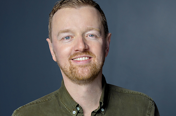 A smiling man with light hair and a beard wearing an olive green button-down shirt against a blue background.