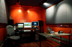 A dark editing studio with red walls, a desk with three monitors, speakers, and a glass table.
