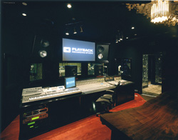 A dark recording studio featuring a large mixing desk, computer monitors, and studio speakers.