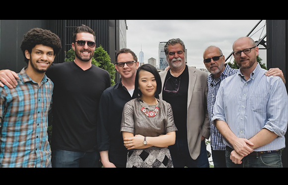 Seven diverse people smiling and posing for a group photo against a city skyline background.