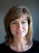 A woman with light brown hair and a black shirt smiling against a gray background.