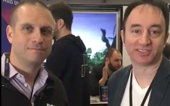 Two men smiling at the camera in an indoor setting with a person in the background.
