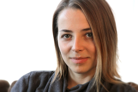 A close-up portrait of a woman with light brown hair looking toward the camera against a white background.