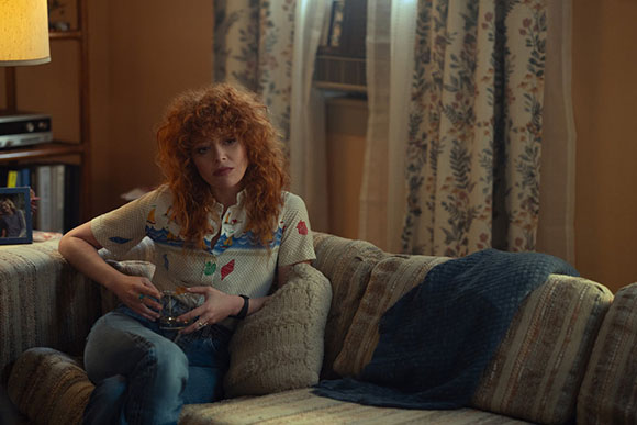 A woman with curly red hair sits on a patterned couch holding a glass in a dimly lit room.