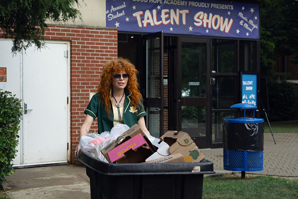 A woman with voluminous red hair and sunglasses stands behind a bin full of boxes in front of a talent show entrance.