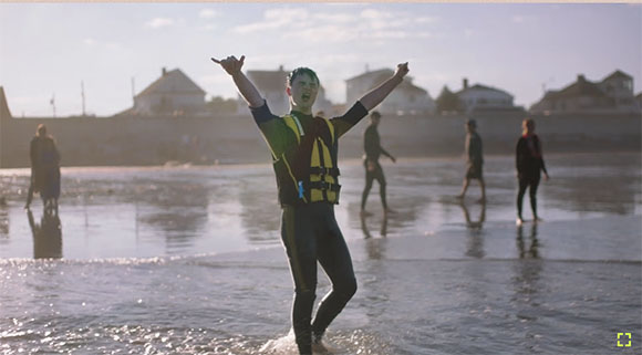 A man in a wetsuit and life vest raises his arms joyfully on a beach with people in the background.