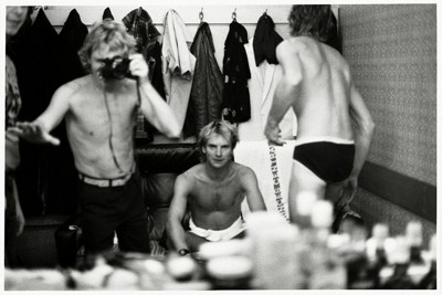 Black and white photo of shirtless men in a dressing room with clothes hanging in the background.