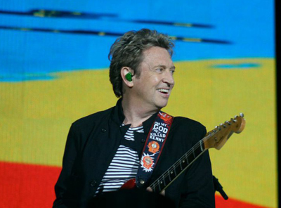 A man in a black jacket and striped shirt smiles while holding an electric guitar on a stage.