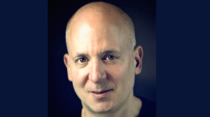 A close-up portrait of a bald man looking directly at the camera against a dark blue background.