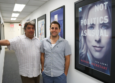 Two men smiling in a hallway next to a framed poster for the show Madam Secretary.