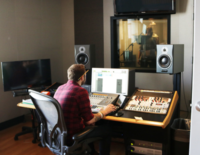 A man in a red plaid shirt sits at a desk with studio monitors, a mixing console, and computer screens.