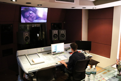 A person sits at a large audio mixing console in a studio with monitors and speakers.