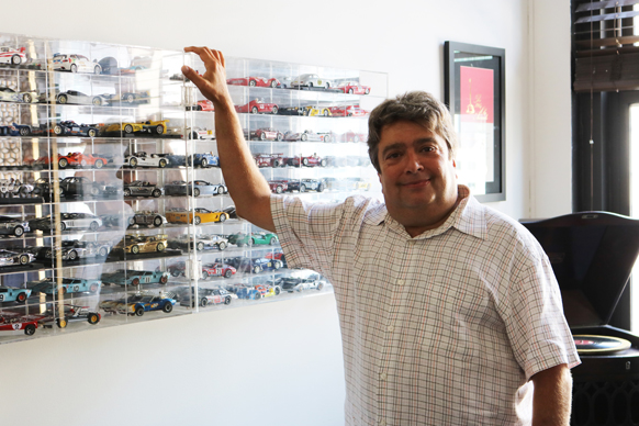 A man in a checkered shirt stands beside a large clear display case filled with numerous miniature model cars.