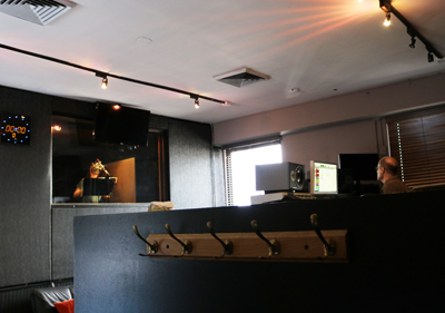 A musician plays a brass instrument in a soundproof booth while a technician works at a desk in a studio.