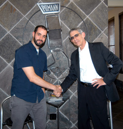 Two men shake hands in front of a large stone wall featuring a Pomann logo.