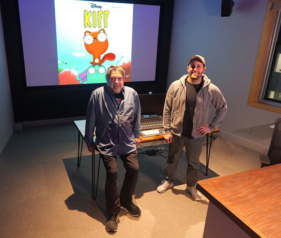 Two men stand in a room before a large screen displaying the Disney animated series Kiff.