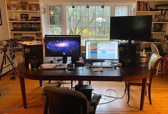 A wooden desk holds two computer monitors, studio speakers, a keyboard, and various cables in a room with a large window.