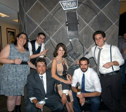 Six people in formal attire posing together in front of a dark stone wall with a decorative logo.