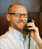 A man with glasses and a beard smiling while holding a black telephone receiver to his ear.